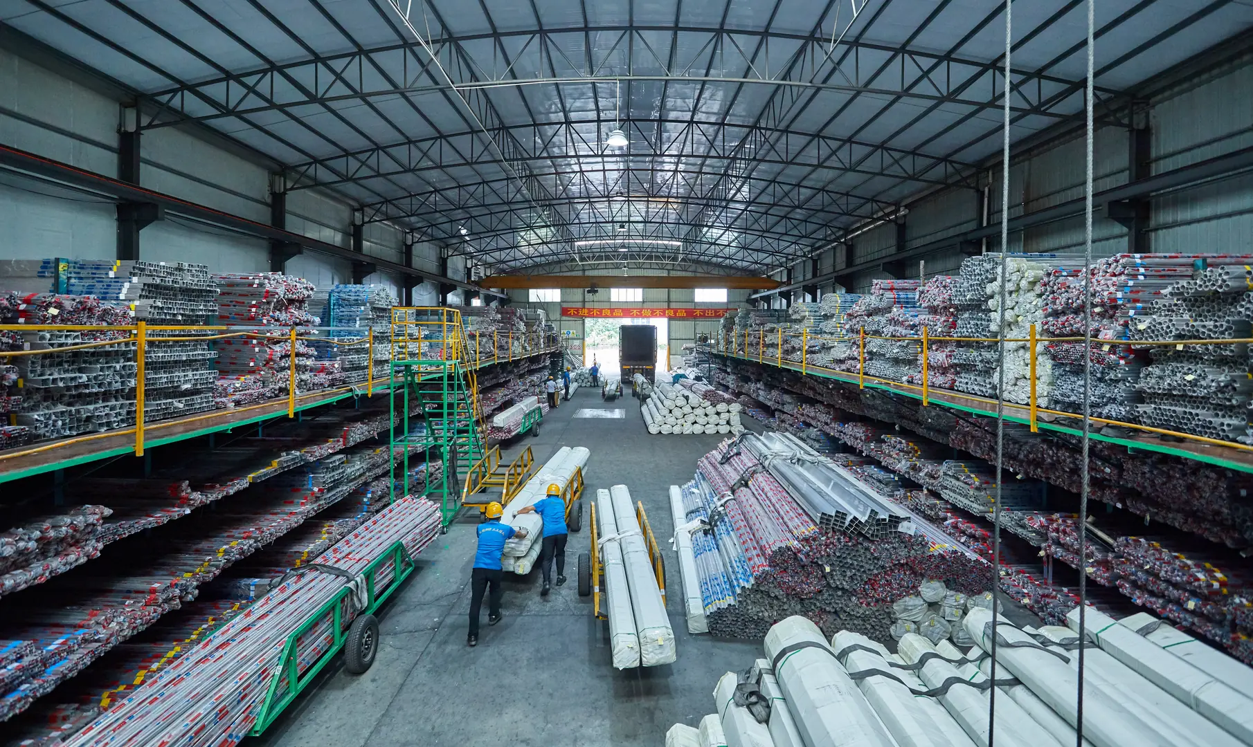 Interior view of a large warehouse filled with organized stacks of stainless steel pipes and workers handling materials